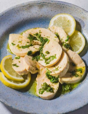 A close up of a sliced poached chicken breast topped with a lemon herb dressing on a plate with lemon slices.