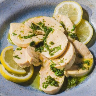 A close up of a sliced poached chicken breast topped with a lemon herb dressing on a plate with lemon slices.