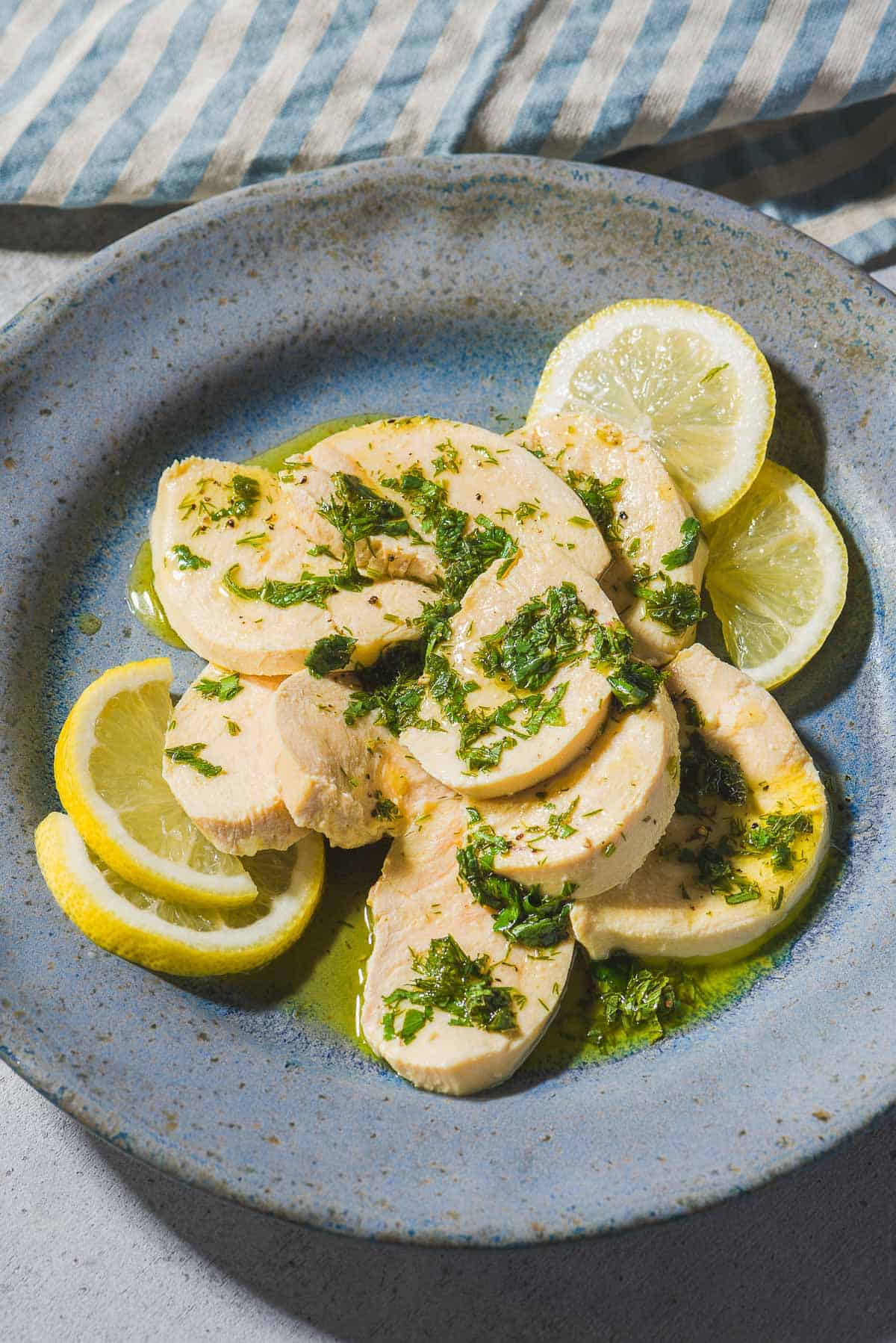 A close up of a sliced poached chicken breast topped with a lemon herb dressing on a plate with lemon slices. Next to this is a cloth napkin.