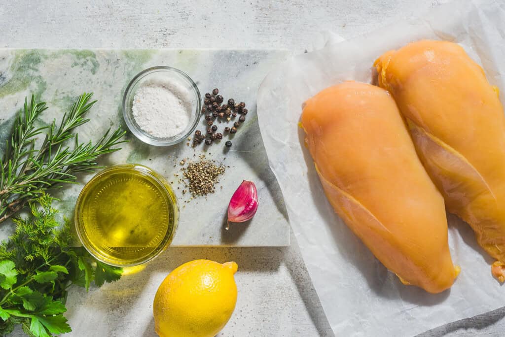 Ingredients for poached chicken breasts with lemon herb dressing including 2 chicken breasts, salt, peppercorns, garlic, sprigs of thyme, dill and parsley, a lemon, olive oil, and black pepper.