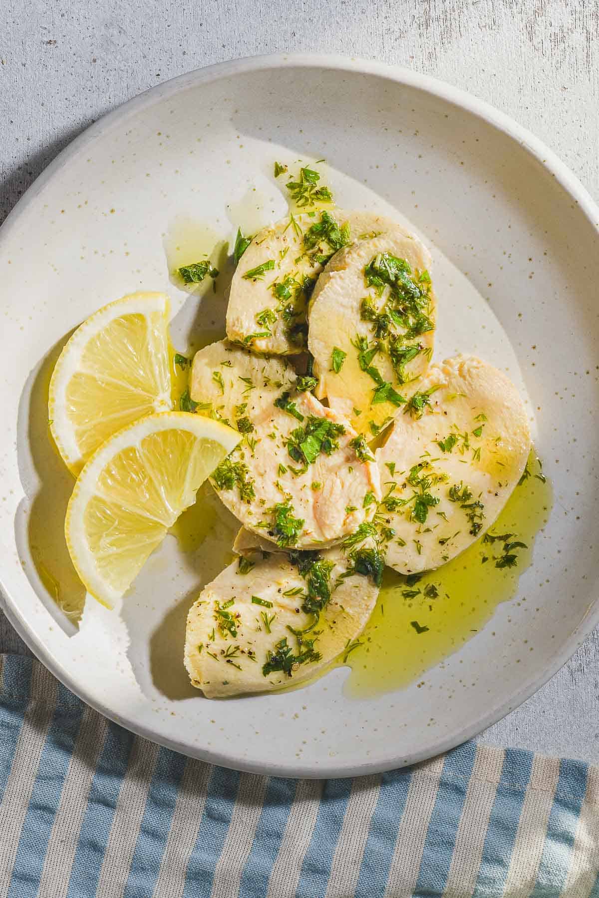 An overhead photo of a sliced poached chicken breast topped with a lemon herb dressing on a plate with lemon slices. Next to this is a cloth napkin.