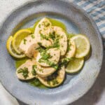 An overhead photo of a sliced poached chicken breast topped with a lemon herb dressing on a plate with lemon slices. Next to this is a cloth napkin.