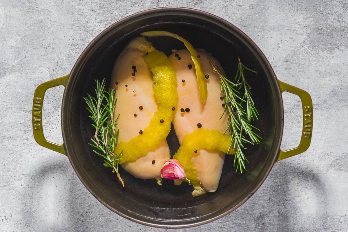 Chicken breasts being poached in water seasoned with salt, peppercorns, garlic, lemon zest and fresh herbs.