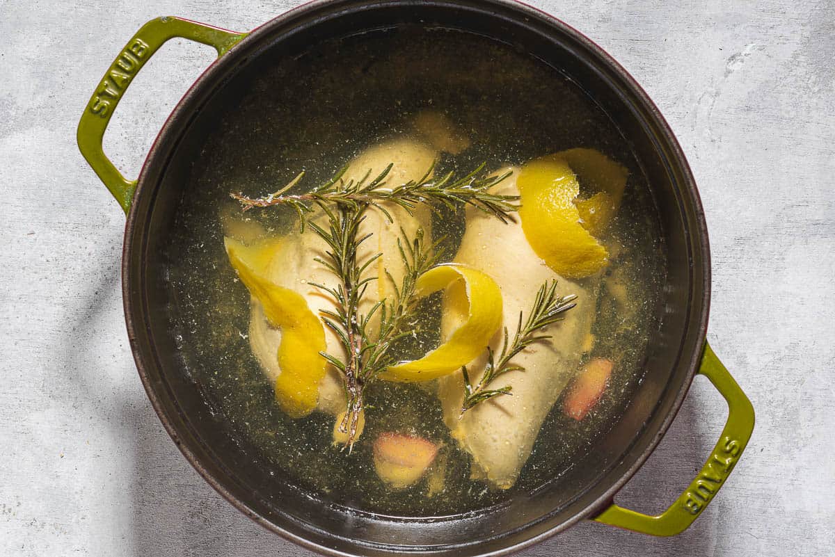 Chicken breasts being poached in water seasoned with salt, peppercorns, garlic, lemon zest and fresh herbs.