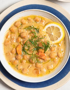 An overhead photo of revithosoupa Greek chickpea soup garnished with dill, a drizzle of olive oil and a lemon slice in a bowl resting on a plate.