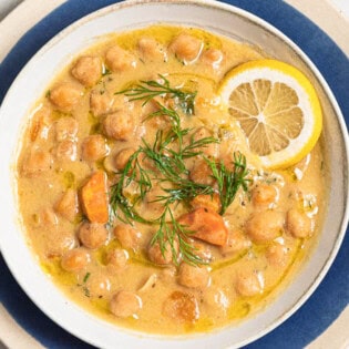 An overhead photo of revithosoupa Greek chickpea soup garnished with dill, a drizzle of olive oil and a lemon slice in a bowl resting on a plate.