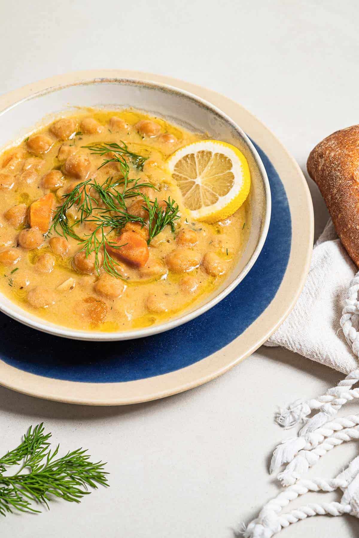 A close up of revithosoupa Greek chickpea soup garnished with dill, a drizzle of olive oil and a lemon slice in a bowl resting on a plate.