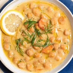 A close up of revithosoupa Greek chickpea soup garnished with dill, a drizzle of olive oil and a lemon slice in a bowl resting on a plate.