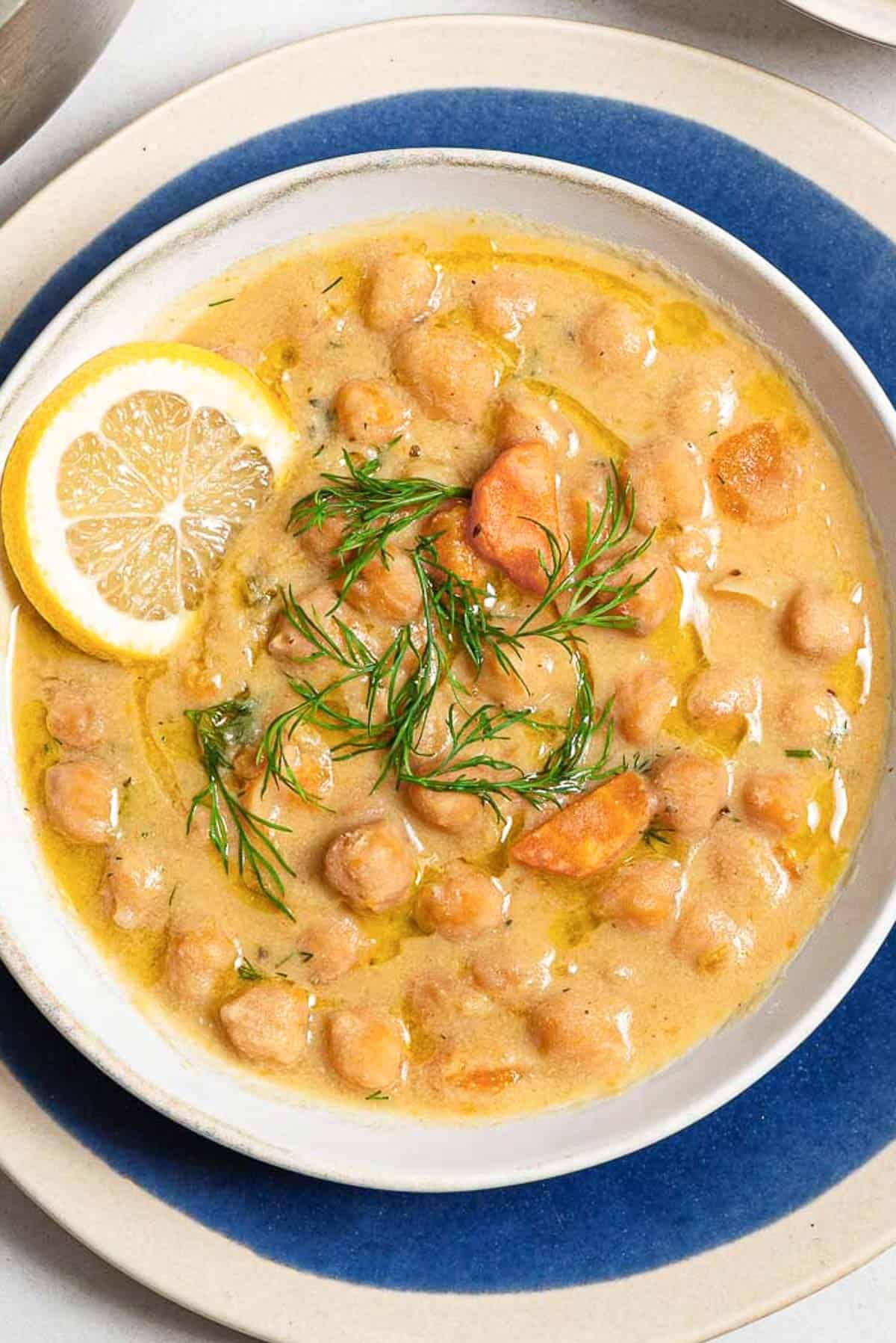 A close up of revithosoupa Greek chickpea soup garnished with dill, a drizzle of olive oil and a lemon slice in a bowl resting on a plate.