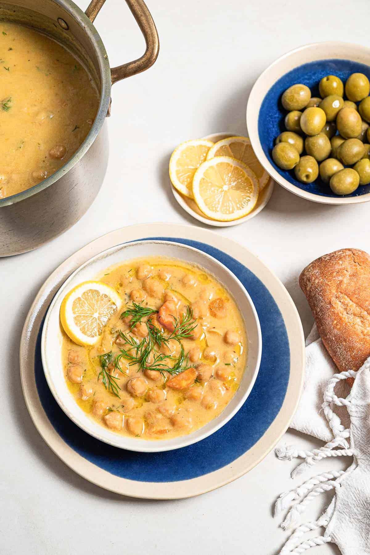 Revithosoupa Greek chickpea soup garnished with dill, a drizzle of olive oil and a lemon slice in a bowl resting on a plate. Next to this is a baguette wrapped in a towel, a bowl of green olives, a plate of lemon slices, and the rest of the soup in a large pot.