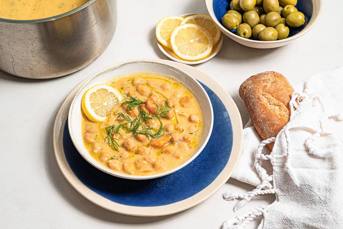 A close up of revithosoupa Greek chickpea soup garnished with dill, a drizzle of olive oil and a lemon slice in a bowl resting on a plate. Next to this is a baguette wrapped in a towel, a bowl of green olives, a plate of lemon slices, and the rest of the soup in a large pot.