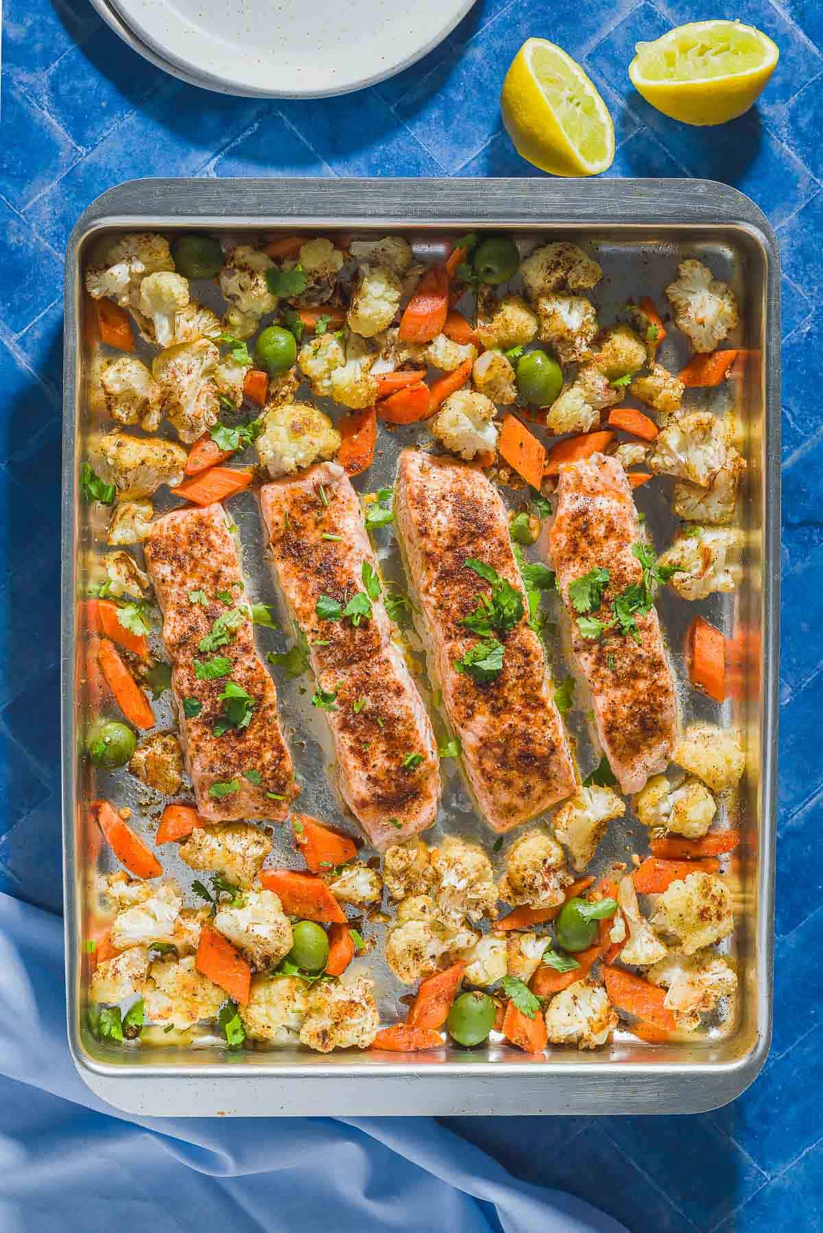 An overhead photo of baharat roasted salmon and vegetables on a sheet pan. Next to this is a stack of 2 plates, 2 juiced lemon halves, and a cloth napkin.