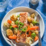 An overhead photo of a baharat roasted salmon fillet on a bed of vegetables and olives on a plate with a fork and knife. Next to this is a glass of water and salt and pepper shakers.