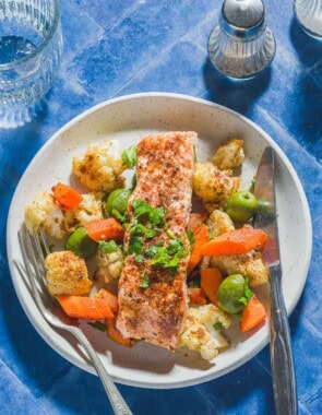 An overhead photo of a baharat roasted salmon fillet on a bed of vegetables and olives on a plate with a fork and knife. Next to this is a glass of water and salt and pepper shakers.