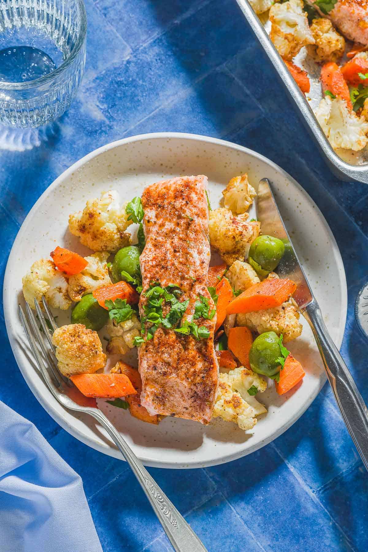 An overhead photo of a baharat roasted salmon fillet on a bed of vegetables and olives on a plate with a fork and knife. Next to this is a glass of water and the rest of the roasted salmon recipe on a sheet pan.