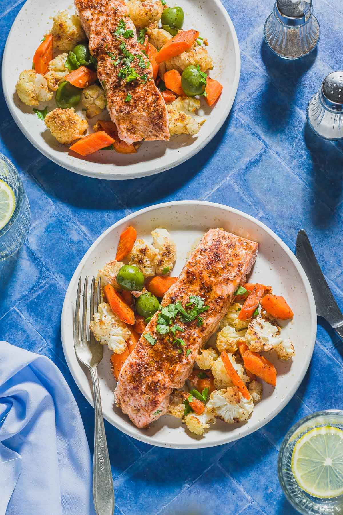 An overhead photo of a 2 servings of the baharat roasted salmon and vegetables on plates, one with a fork. Next to these are 2 glasses of water with lemon wheels, a knife, a cloth napkin, and salt and pepper shakers.