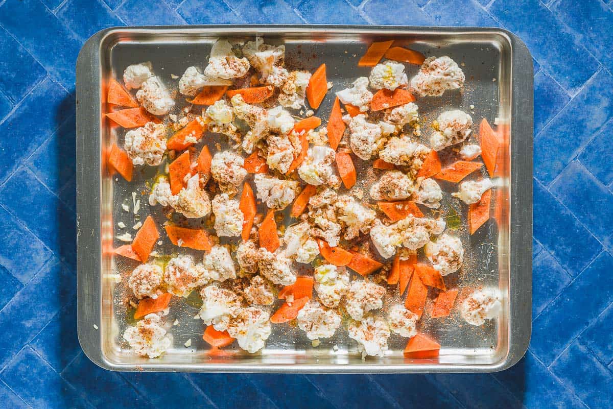 An overhead photo of uncooked carrot pieces and cauliflower florets seasoned with spices and drizzled with olive oil on a sheet pan.