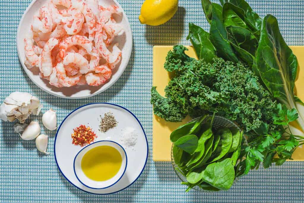 Ingredients for pan seared shrimp including peeled and deveined shrimp, a lemon, garlic, cooking greens, salt, black pepper, red pepper flakes, and olive oil.