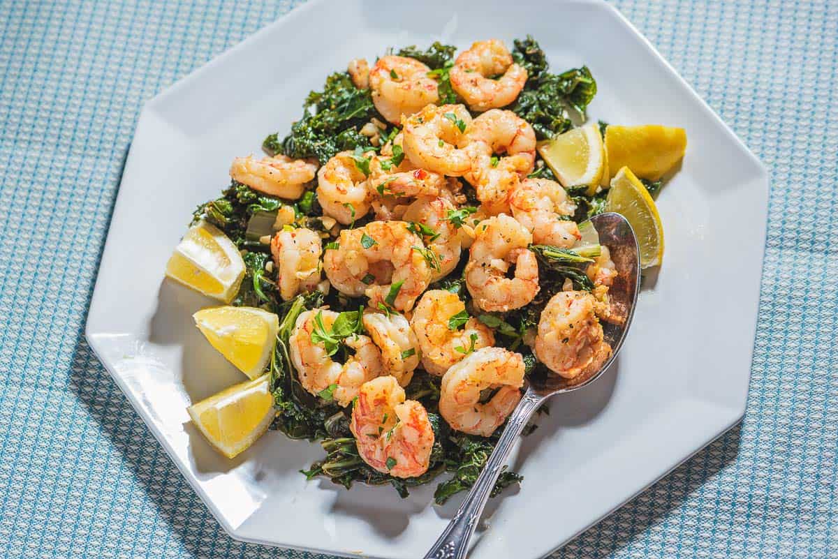 A close up of pan seared shrimp over braised lemon and garlic greens on a plate with lemon wedges and a spoon.