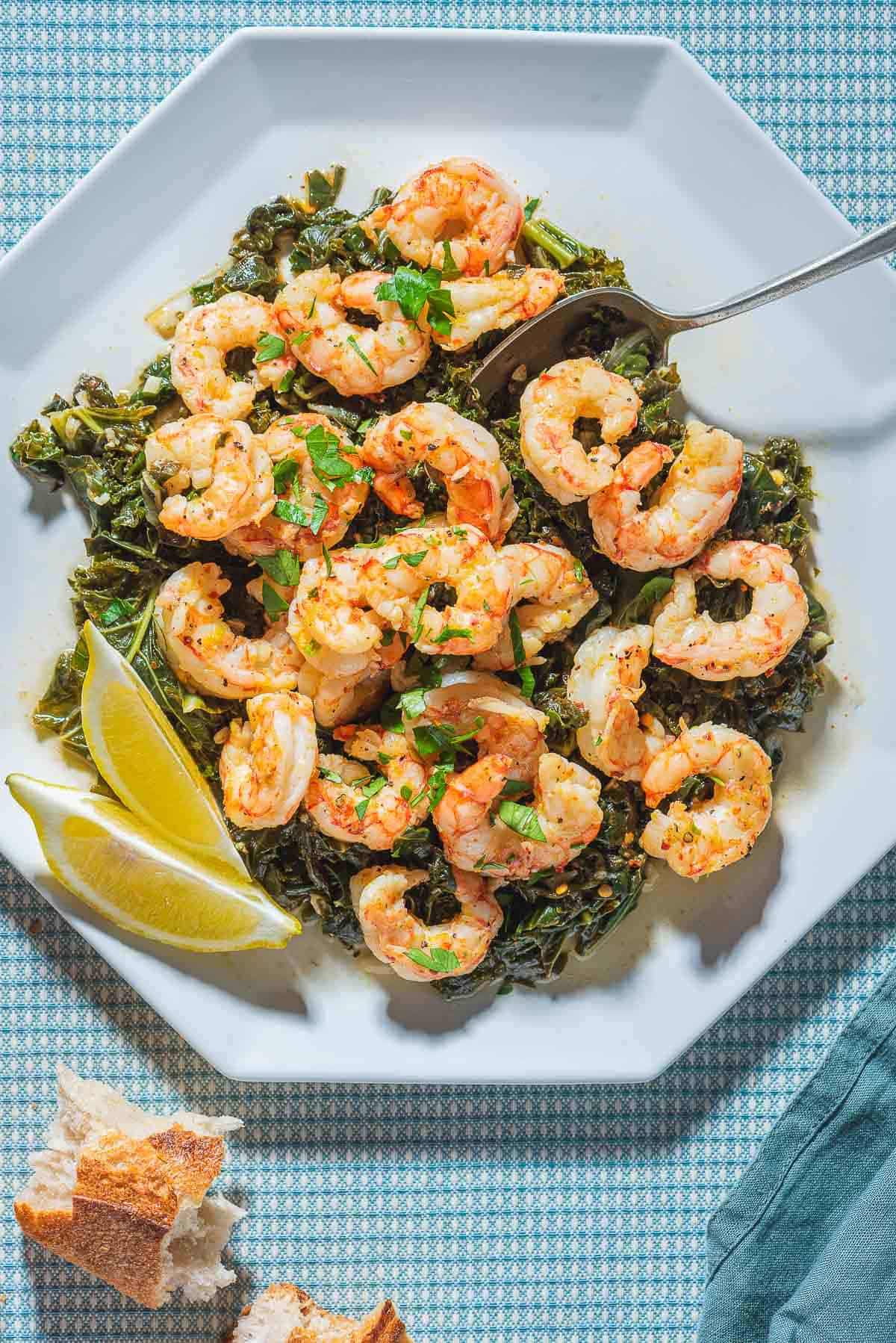 An overhead photo of pan seared shrimp over braised lemon and garlic greens on a plate with lemon wedges and a spoon. Next to this is a cloth napkin and some crusty bread.