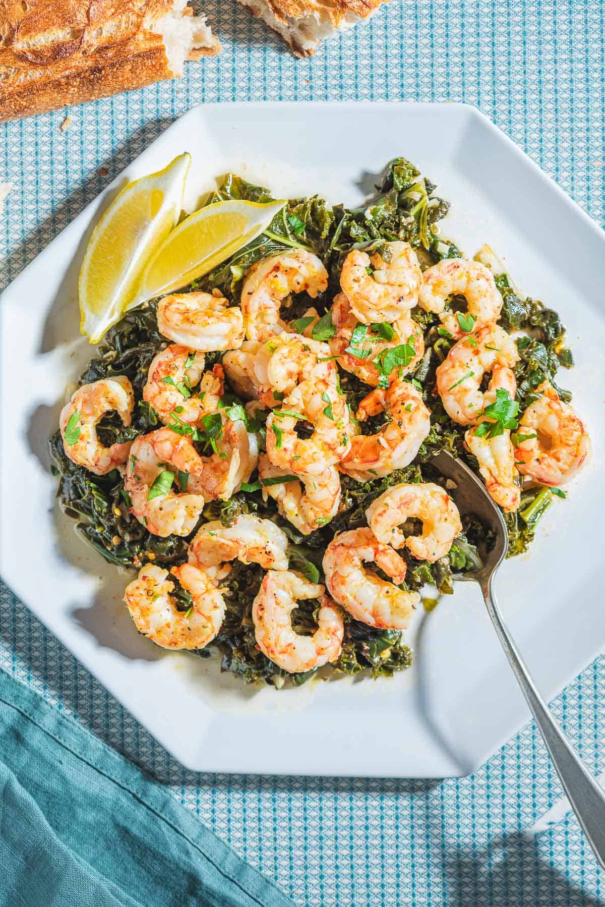 An overhead photo of pan seared healthy shrimp recipe over braised lemon and garlic greens on a white plate with lemon wedges and a spoon. Next to this is a cloth napkin and some crusty bread.