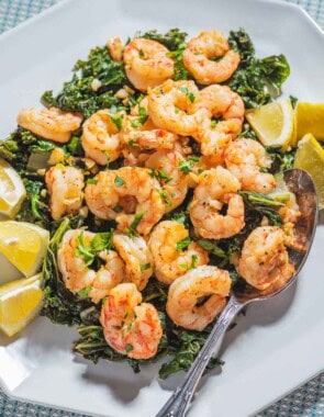 A close up of pan seared shrimp over braised lemon and garlic greens on a plate with lemon wedges and a spoon.