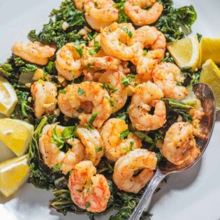 A close up of pan seared shrimp over braised lemon and garlic greens on a plate with lemon wedges and a spoon.