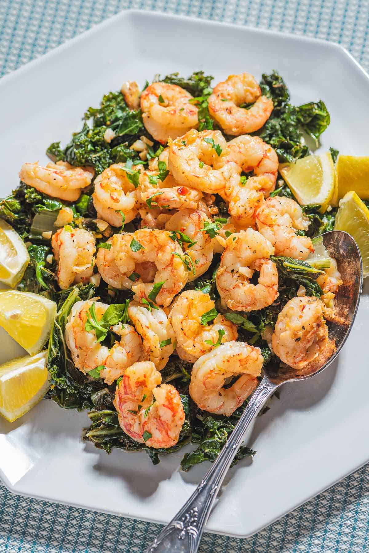 A close up of pan seared shrimp over braised lemon and garlic greens on a plate with lemon wedges and a spoon.