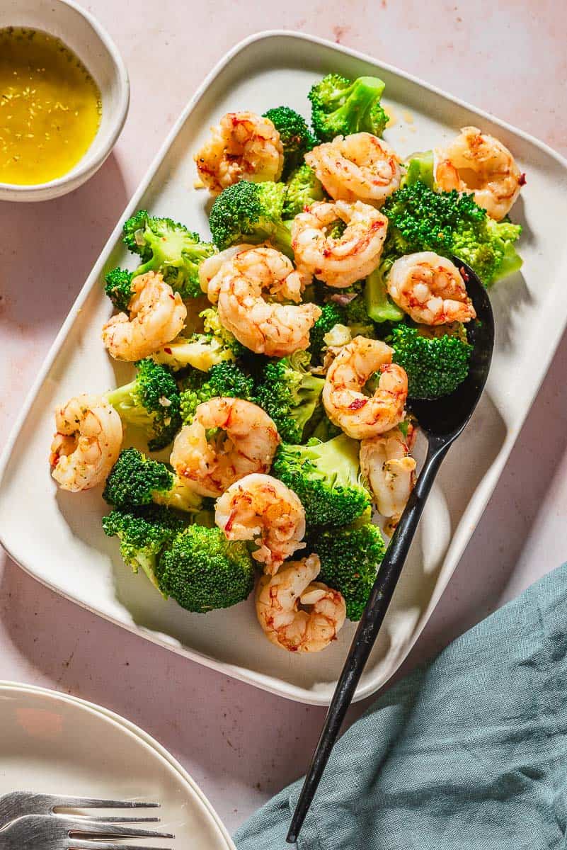 An overhead photo of the cooked shrimp and broccoli recipe on a platter with a serving spoon. Next to this is a stack of 2 plates with 2 forks, a cloth napkin, and a bowl of the ladolemono sauce.