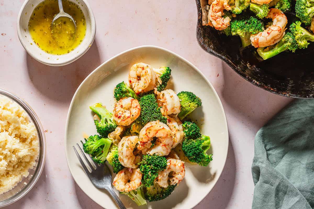 An overhead photo of the cooked shrimp and broccoli recipe on a plate with a fork. Next to this is a cloth napkin, a bowl of ladolemono sauce in a bowl with a spoon, a bowl of couscous, and the rest of the shrimp and broccoli recipe in a skillet.