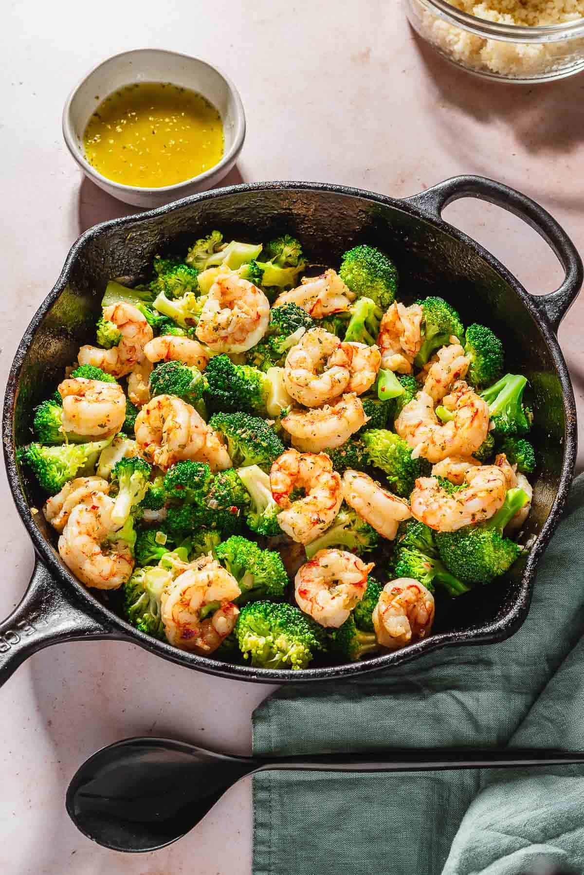 An overhead photo of the cooked shrimp and broccoli skillet. Next to this is a cloth napkin, a serving spoon, a bowl of couscous, and a bowl of ladolemono sauce.