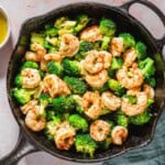 An overhead photo of the cooked shrimp and broccoli skillet. Next to this is a cloth napkin, a bowl of couscous, and a bowl of ladolemono sauce.