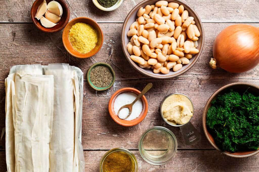 Ingredients for vegan skillet spanakopita including white beans, hummus, olive oil, green olive brine, nutritional yeast, dried dill, salt, black pepper, frozen spinach, onion, garlic and phyllo dough.