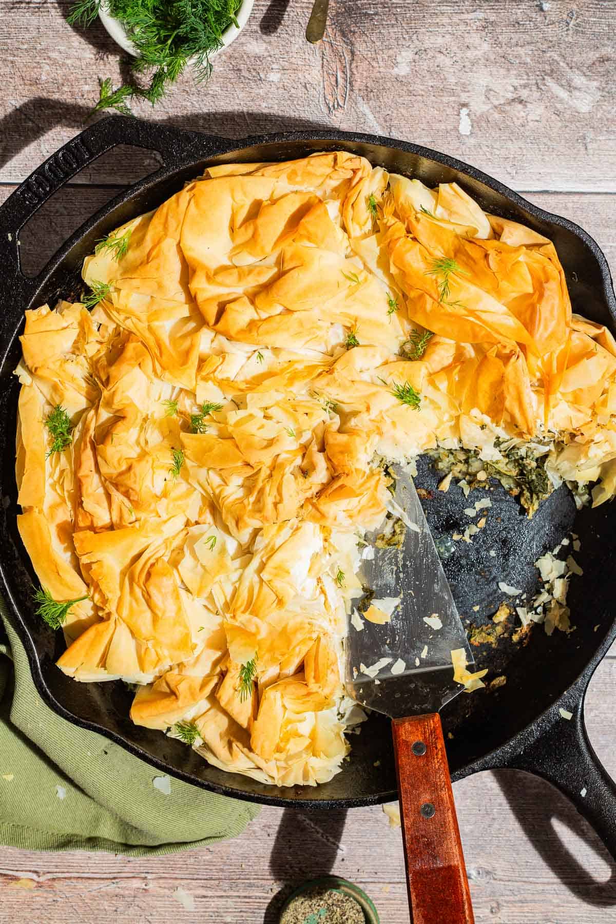 An overhead photo of vegan skillet spanakopita in a skillet with a few pieces removed. Next to this are bowls pepper and dill and a cloth napkin.