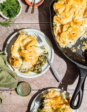 An overhead photo of 2 servings of vegan skillet spanakopita on plates with forks, the rest of the spanakopita in a skillet, bowls of dill, salt and pepper and a cloth napkin.