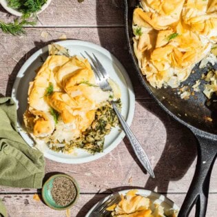 An overhead photo of 2 servings of vegan skillet spanakopita on plates with forks, the rest of the spanakopita in a skillet, bowls of dill, salt and pepper and a cloth napkin.