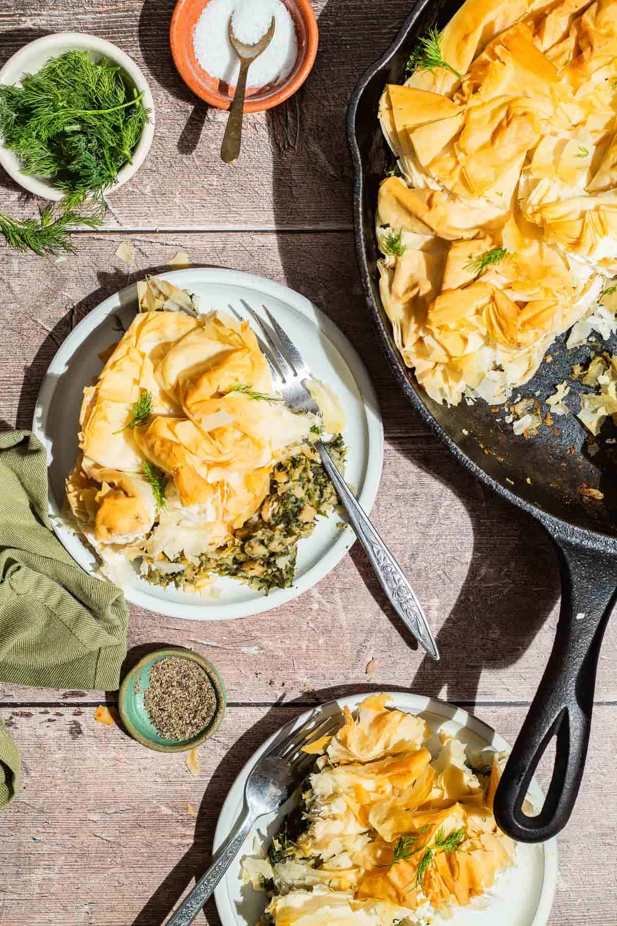 An overhead photo of 2 servings of vegan skillet spanakopita on plates with forks, the rest of the spanakopita in a skillet, bowls of dill, salt and pepper and a cloth napkin.