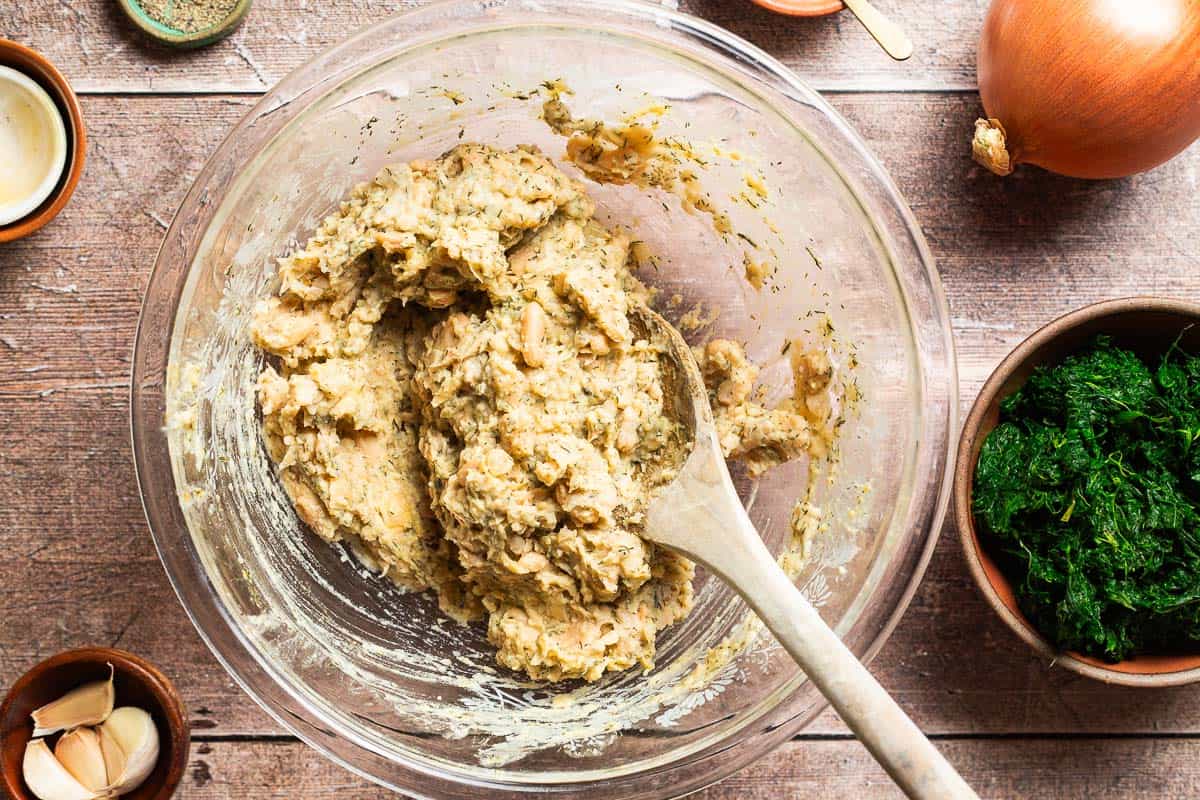 An overhead photo of the filling for the vegan skillet spanakopita just before the spinach is added in a bowl with a wooden spoon. Next to this is a bowl of garlic cloves, a bowl of frozen spinach, and an onion.