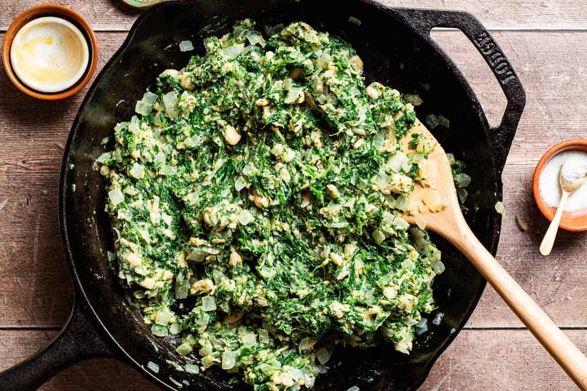 An overhead photo of the filling for the vegan skillet spanakopita in a skillet with a wooden spoon. Next to this is a bowl of salt and a stack of empty bowls.