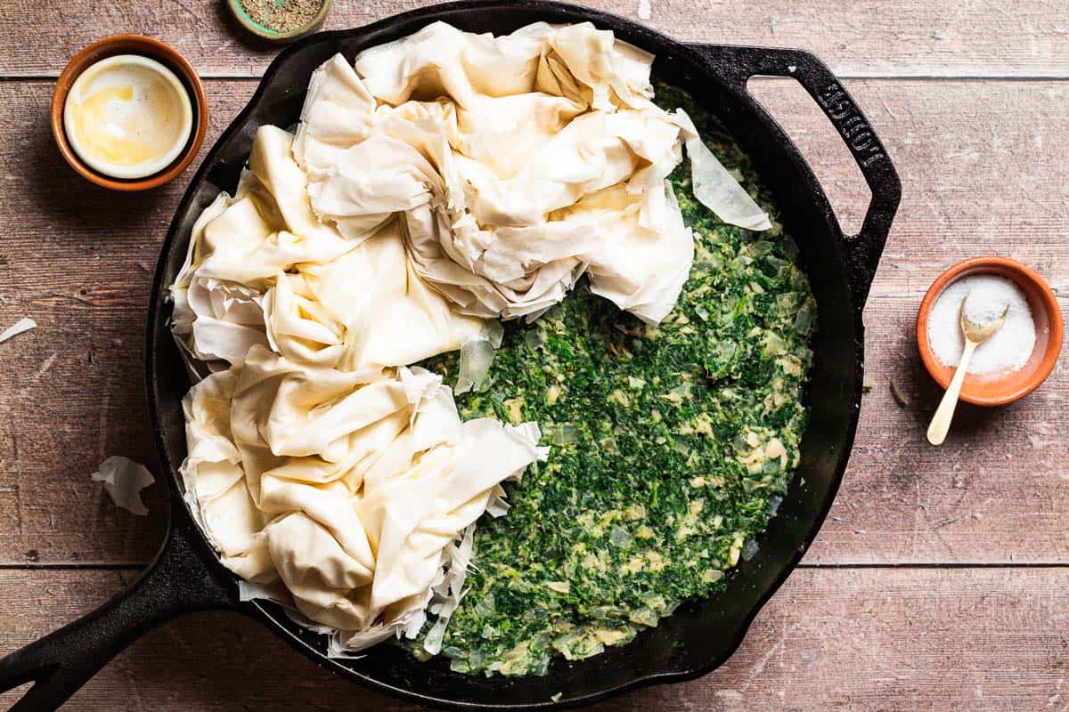 An overhead photo of phyllo dough being added to the top of the uncooked vegan skillet spanakopita in a skillet. Next to this are bowls of salt and pepper, and a stack of empty bowls.