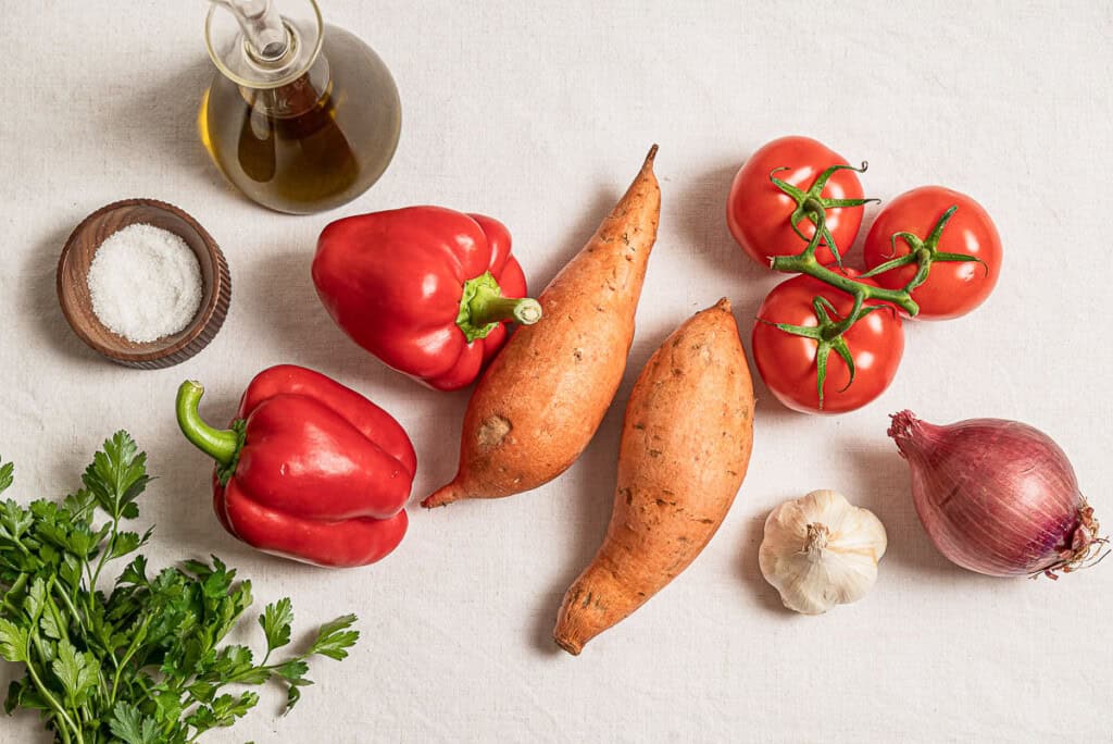 Ingredients for sweet potato soup including red bell peppers, sweet potatoes, red onion, garlic, tomatoes, olive oil, salt, and parsley.