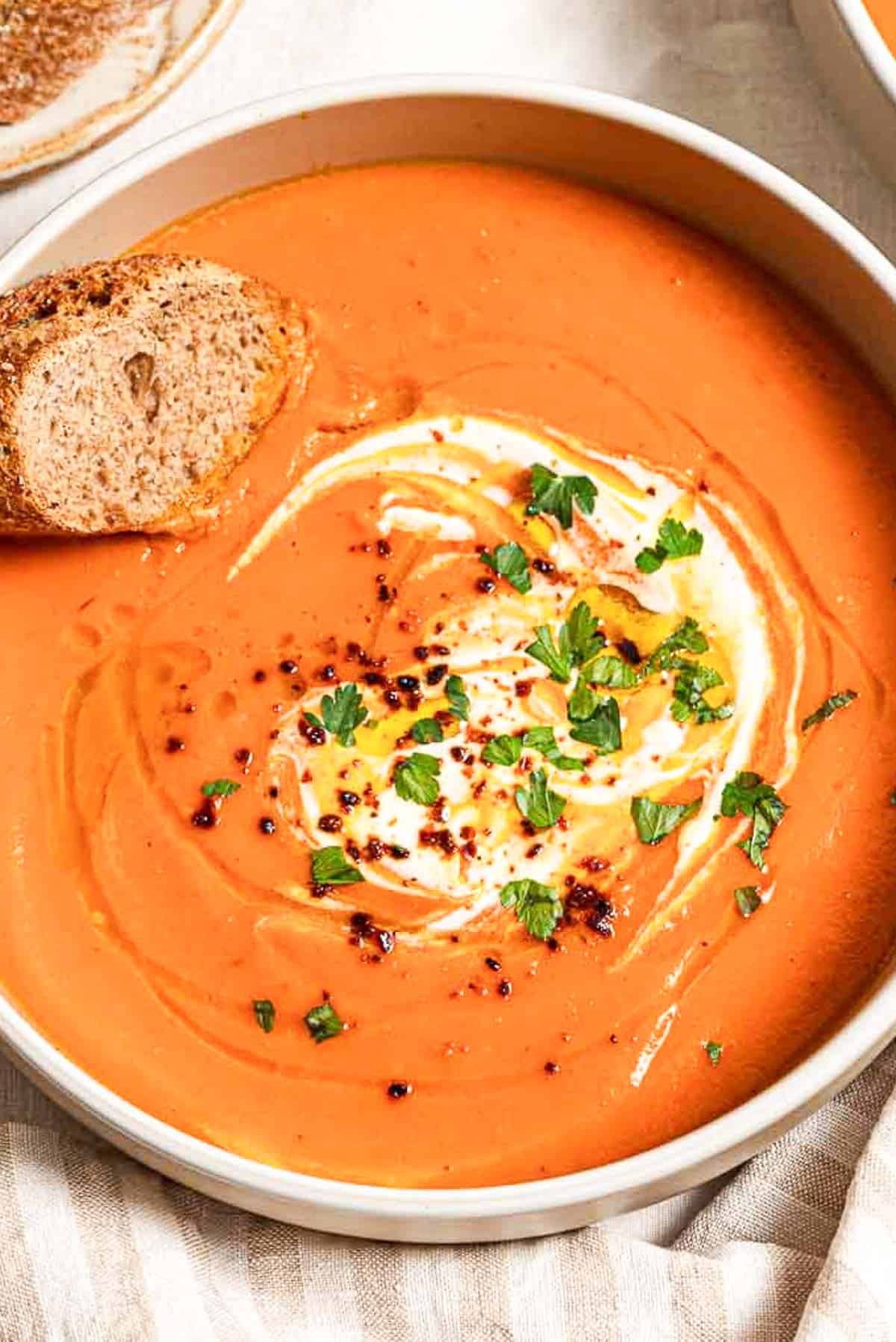 A close up of sweet potato soup topped with a dollop of greek yogurt, a drizzle of olive oil, Aleppo pepper and chopped parsley in a bowl along with a slice of bread. Next to this is a cloth napkin.