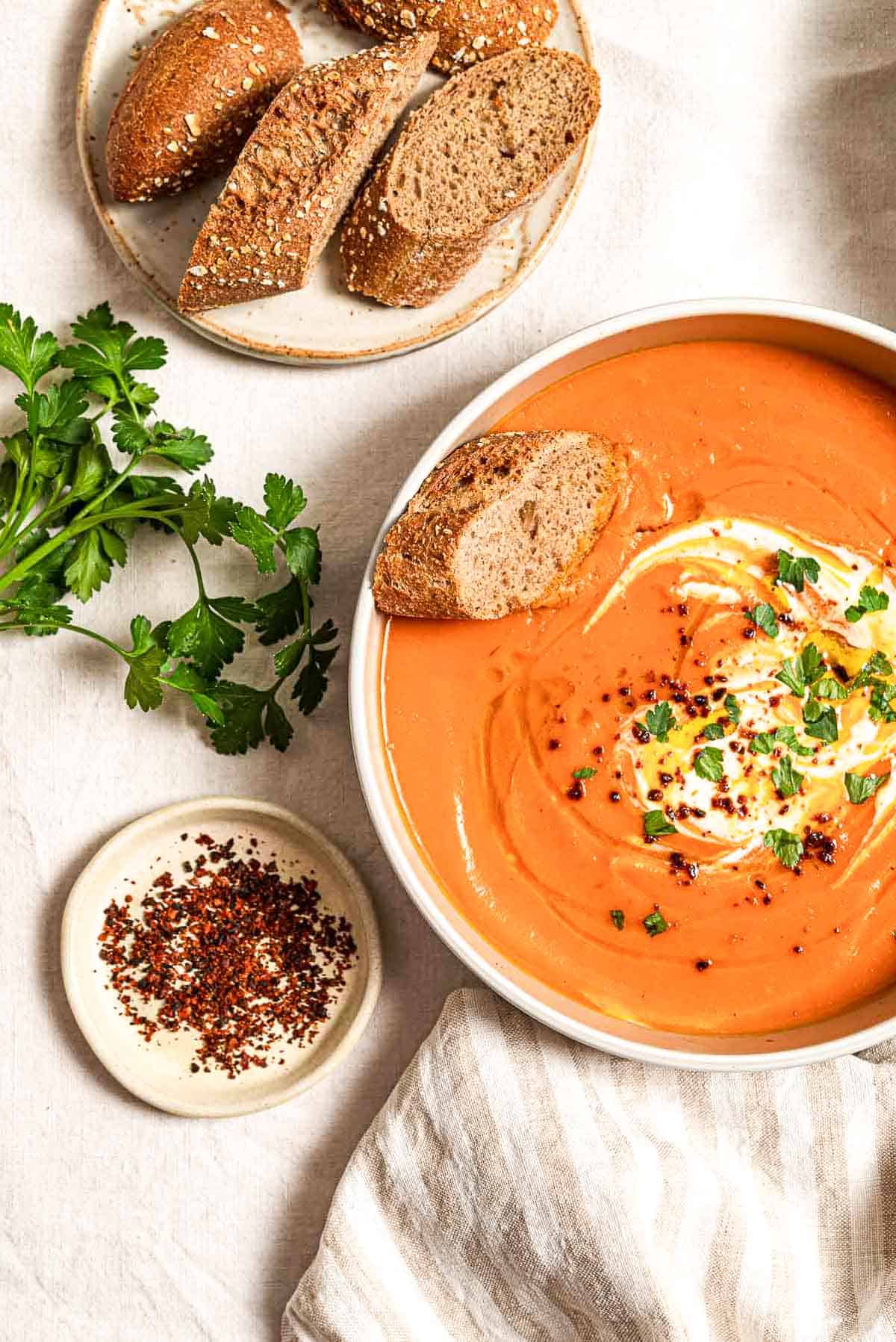 An overhead photo of sweet potato soup topped with a dollop of greek yogurt, a drizzle of olive oil, Aleppo pepper and chopped parsley in a bowl along with a slice of bread. Next to this is more sliced bread on a plate, a few sprigs of parsley, a small bowl of aleppo pepper and a cloth napkin.