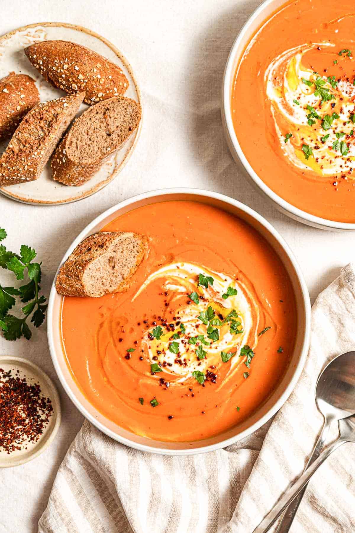 An overhead photo of 2 bowls of sweet potato soup, each topped with a dollop of greek yogurt, a drizzle of olive oil, Aleppo pepper and chopped parsley. Next to these is a plate with more sliced bread, a few sprigs of parsley, a small bowl of aleppo pepper and a cloth napkin with two spoons.