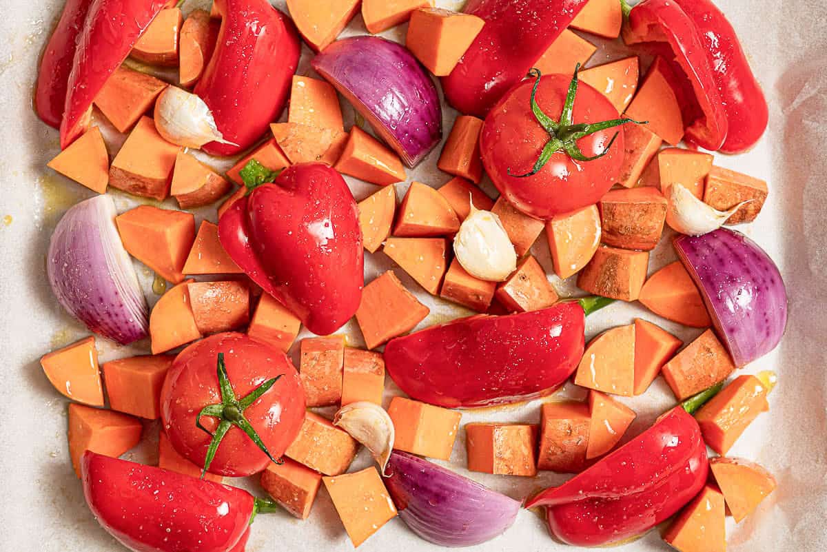 An overhead photo of the uncooked, cut vegetables for the sweet potato soup recipe, drizzled with olive oil, on a parchment lined sheet pan.