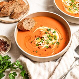 Two bowls of sweet potato soup, each topped with a dollop of greek yogurt, a drizzle of olive oil, Aleppo pepper and chopped parsley. Next to these is a plate with more sliced bread, a few sprigs of parsley, a small bowl of aleppo pepper and a cloth napkin with two spoons.