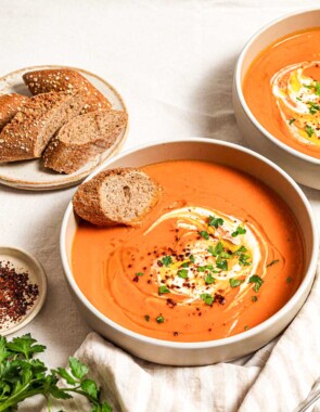 Two bowls of sweet potato soup, each topped with a dollop of greek yogurt, a drizzle of olive oil, Aleppo pepper and chopped parsley. Next to these is a plate with more sliced bread, a few sprigs of parsley, a small bowl of aleppo pepper and a cloth napkin with two spoons.