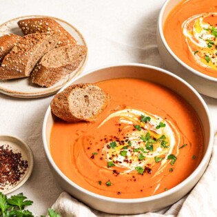 Two bowls of sweet potato soup, each topped with a dollop of greek yogurt, a drizzle of olive oil, Aleppo pepper and chopped parsley. Next to these is a plate with more sliced bread, a few sprigs of parsley, a small bowl of aleppo pepper and a cloth napkin with two spoons.