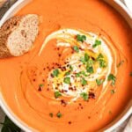 An overhead photo of sweet potato soup topped with a dollop of greek yogurt, a drizzle of olive oil, Aleppo pepper and chopped parsley in a bowl along with a slice of bread. Next to this is a cloth napkin.