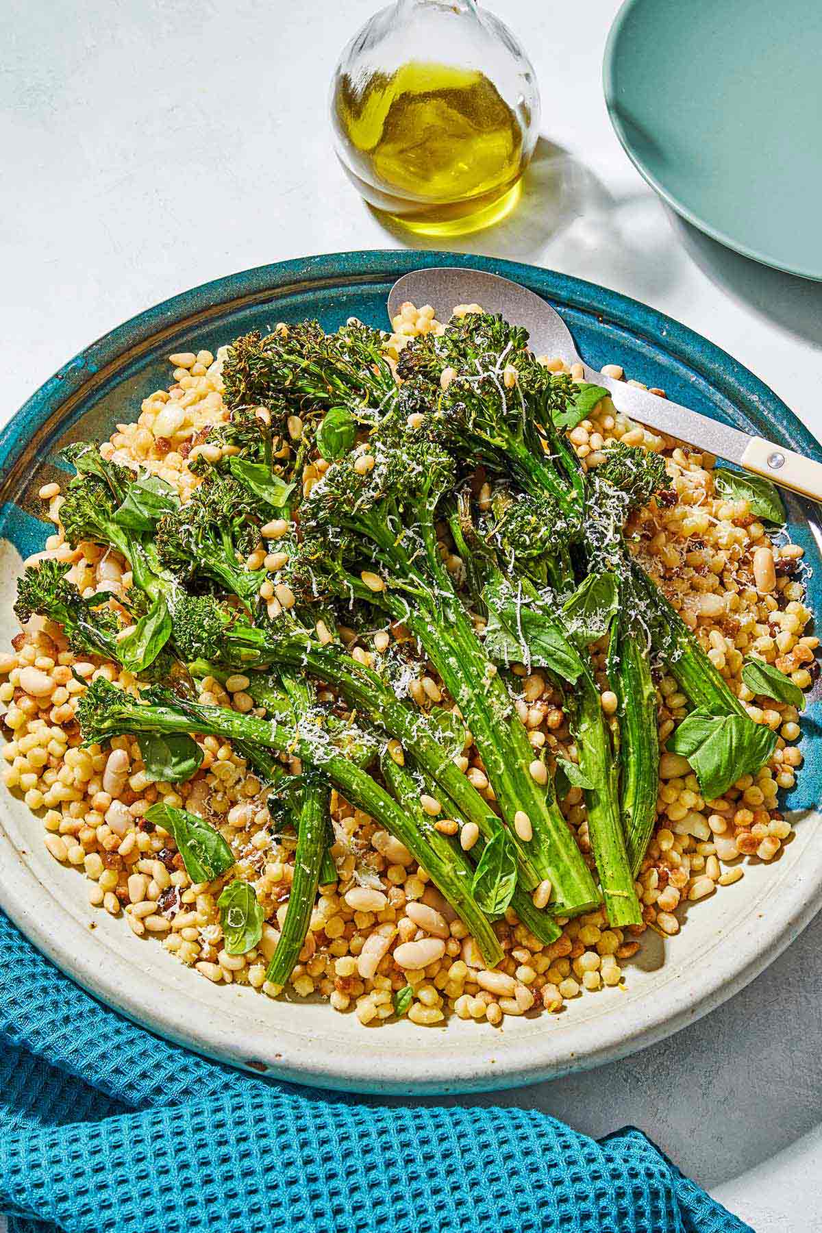 Fregola sarda with broccolini on a platter with a spoon. Next to this is a cloth napkin, a small jug of olive oil and an empty plate.